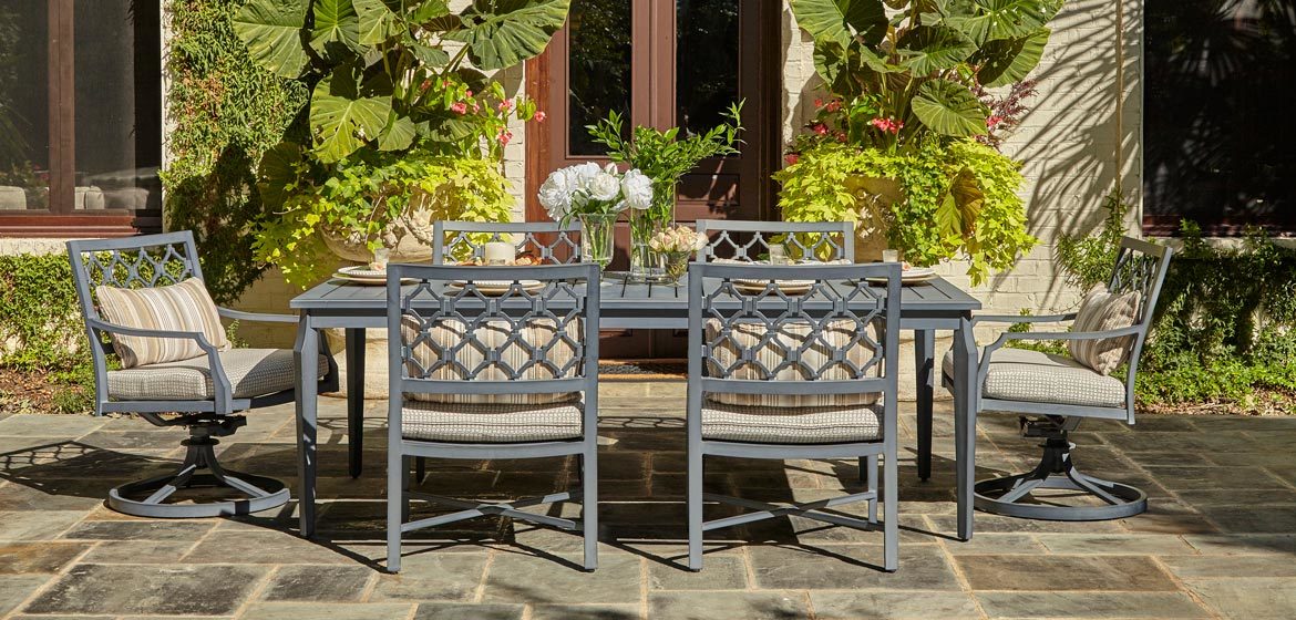 Klaussner Outdoor Mirage collection rectangular dining table with swivel rockers and side chairs on a flagstone patio in front of a painted brick home