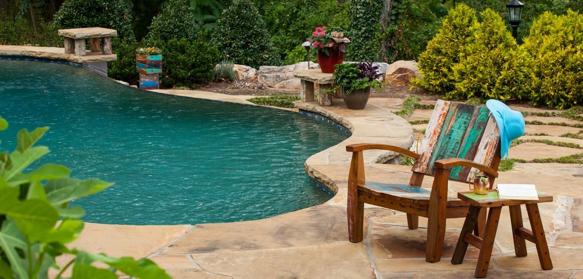 Warehouse 2120's recycled boatwood outdoor lounge chair and side table on an irregular flagstone patio by a beautiful pool with a diving rock